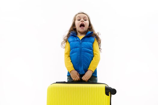 Mischievous Caucasian little child traveler girl, holds a heavy yellow suitcase, expresses surprise, poses with open mouth on white background. Copy space for your advertising text. Tourism and travel