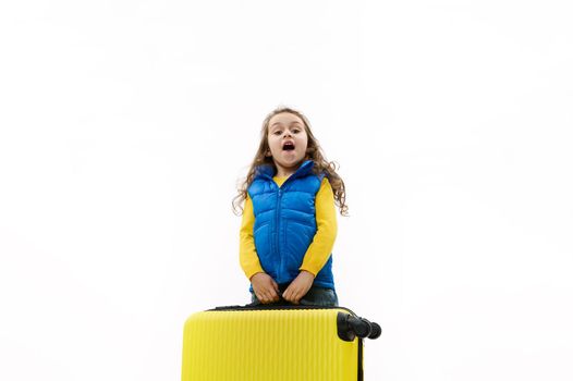 Caucasian beautiful little kid girl in blue jacket, going for weekend getaway, carrying a heavy yellow suitcase, isolated on white background. Travel. Trip. Journey. Air Flight. Copy advertising space