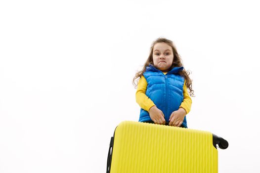 Caucasian mischievous traveler child girl in blue jacket, going for vacations, carrying a heavy yellow valise, isolated on white background. Travel. Weekend Trip. Journey. Air Flight. Copy ad space