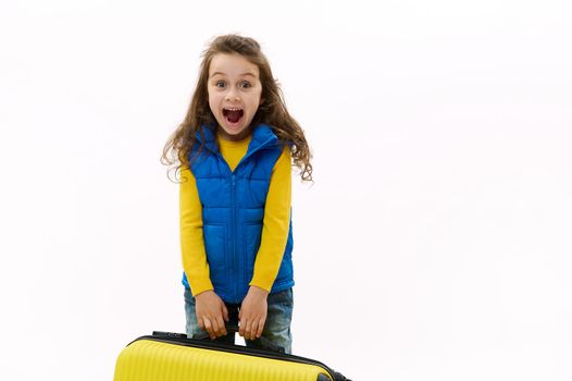 Mischievous little traveler kid girl 5-6 years old, holding yellow suitcase, isolated on white background. Passenger traveling abroad on weekends getaway. Air flight journey concept. Copy ad space