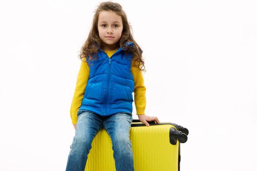 Adorable child girl in blue jacket and jeans, sitting on yellow suitcase, looking at camera, isolated on white background. Charming kid with travel bag, going for weekend getaway. Copy space for ads