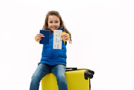 Happy Caucasian little child girl traveler sits on a yellow suitcase, handing a passport and air ticket to camera, isolated on white background. Kid travelling abroad for winter vacations. Copy space