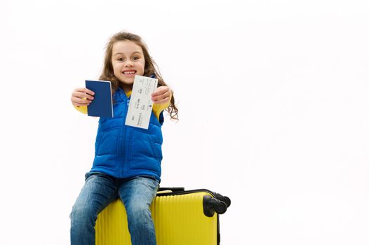 Mischievous overjoyed child girl sitting on yellow suitcase, holding out at camera passport and air ticket, rejoicing at upcoming travel abroad, isolated over white background. Copy advertising space