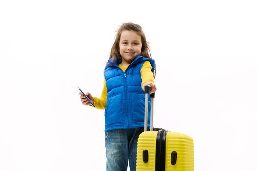 Caucasian lovely little kid girl, wearing a blue jacket, jeans and yellow pullover, smiles looking at camera, posing with a travel bag and boarding pass on white background. Tourism Travel. Copy space