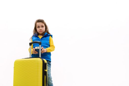 Cute baby girl in blue down jacket with yellow suitcase and boarding pass, going for weekend getaway abroad, isolated over white background with free advertising space. Travel Journey Children concept