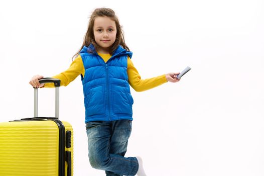 Happy child girl wearing blue denim and jacket with yellow suitcase and boarding pass, looking at camera, isolated on white background with copy advertising space. Kid with baggage going on vacations