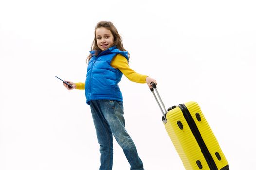 Full length: fashionable little child, traveler girl in blue jeans, yellow shirt and down jacket, traveling abroad for holidays, walking with suitcase on white background. Journey Travel Tourism Trip