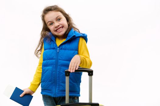 Mischievous European little girl in yellow sweater and blue down jacket, holds boarding pass and suitcase, smiles looking at camera, isolated on white background. Cute child travel abroad for holidays