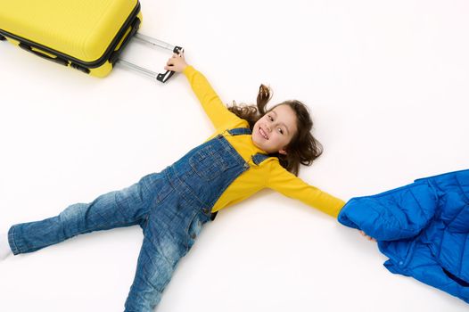Top view cute little child girl tourist in blue denim overalls, with yellow sweater and suitcase, lying on white background, advertising space. Travel abroad. Getaway. Air flight trip journey concept