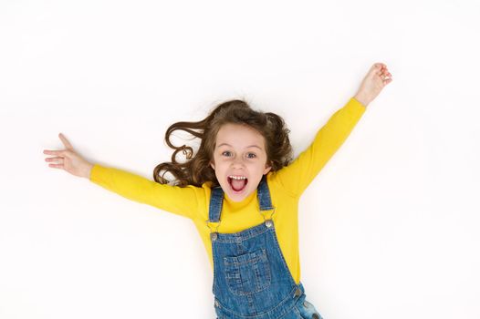 View from above: focus on facial expressions of mischievous Caucasian baby girl with outstretched arms, rejoicing, smiling, looking at camera while lying on white background with copy advertising text