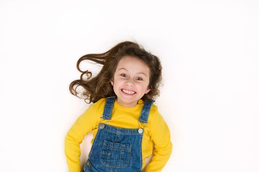 Charming European 5-6 years little child girl, in yellow sweater and blue denim overalls, smiles a cheerful toothy smile, looking at camera, isolated over white background with copy advertising space