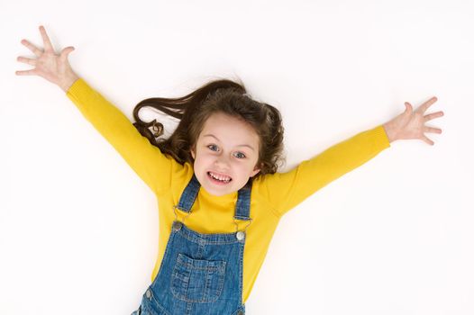 Overhead view of an adorable mischievous Caucasian baby girl with outstretched arms, making faces, rejoicing, smiling, looking at camera while lying on white background with copy advertising text