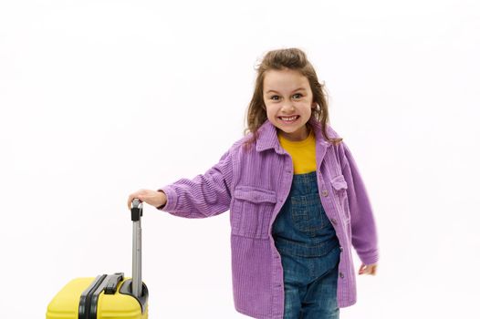 Mischievous traveler child, little girl dressed in purple shirt and denim overalls, going for holidays, posing with yellow suitcase over white background. Kkids travelling abroad. Travel Trip Journey