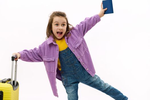 Caucasian adorable mischievous child, a little traveler baby girl in denim overalls, purple shirt, poses with a yellow suitcase and boarding pass over white background. Kids travelling abroad. Tourism
