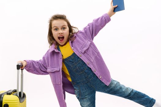 Mischievous European child, lovely traveler little girl wearing purple jacket and blue denim overalls, making faces looking at camera, posing with boarding pass and yellow suitcase on white background