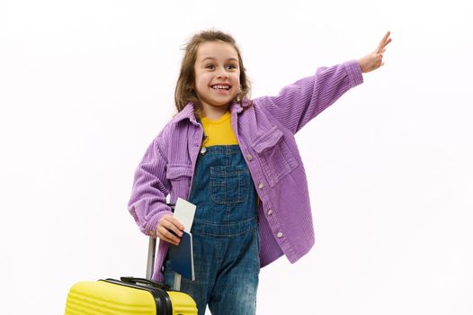 Caucasian happy child girl in violet shirt and blue denim overalls, with boarding pass and yellow suitcase, smiling, waving hello, isolated on white background. Copy space