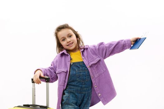 European lovely baby girl in violet velvet jacket, yellow sweater and blue dem nioveralls, going for holidays, smiling a cheerful toothy smile, posing with boarding pass and suitcase, white background