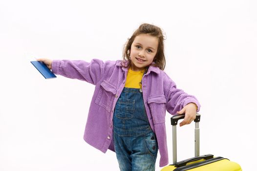 Adorable joyful child girl expressing positive emotions while going for vacations, cutely smiling a cheerful toothy smile at camera, posing with yellow suitcase and boarding pass over white background