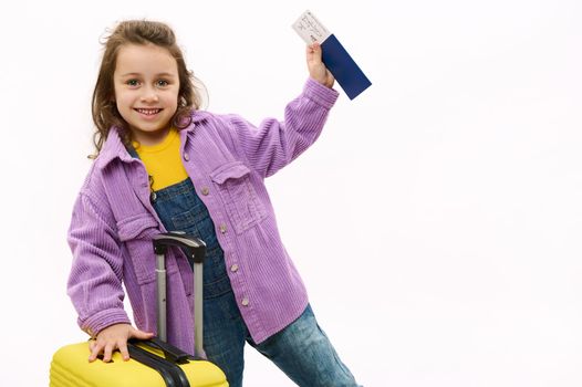 Caucasian adorable little girl, child traveler tourist in purple velvet jacket, going for holidays, cutely smiles looking at camera, posing with yellow suitcase and boarding pass over white background