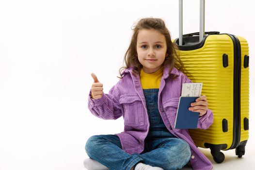 Happy child girl in purple shirt and denim overalls, sitting near a yellow suitcase, holding a boarding pass and showing thumb up to camera, wainting her flight, isolated on white. Kids travel abroad