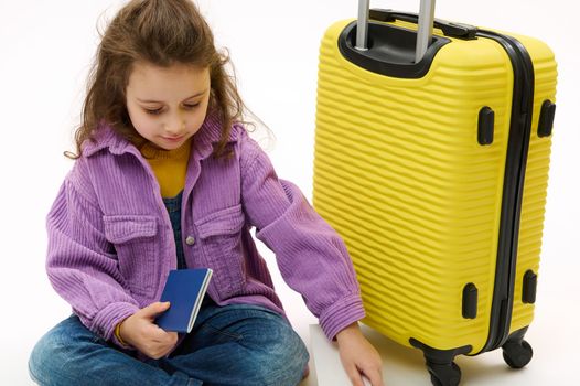 Adorable Caucasian child girl, dressed in violet shirt and denim overalls, a little traveler tourist checking her air ticket before boarding the flight, sitting near yellow suitcase, white background