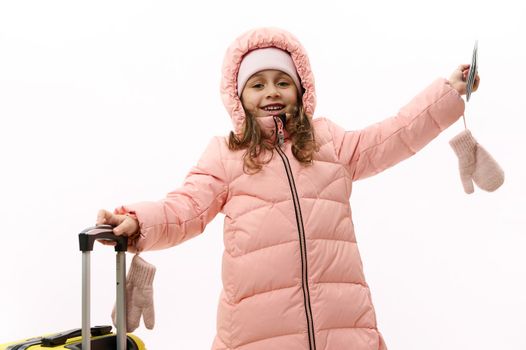 Joyful baby girl in warm pink down clothes, going for winter holidays, posing with boarding pass and a suitcase on white background with free advertising space