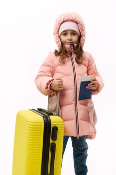 Full-length portrait: Beautiful little girl in pink down coat, going for vacations, looking at camera, posing with suitcase and boarding pass on white background with copy space. Travel Trip Journey
