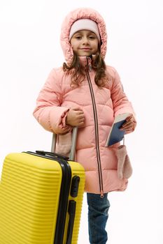 Lovely baby girl in pink warm down coat and blue denim jeans, going on vacations smiles cutely looking at camera, holding boarding pass and flight ticket, posing with yellow suitcase, white background