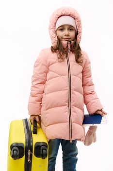 Adorable Caucasian baby girl wearing pink down coat, going for winter holidays, travelling abroad, holding boarding pass and yellow suitcase, smiling looking at camera over white backdrop. Copy space