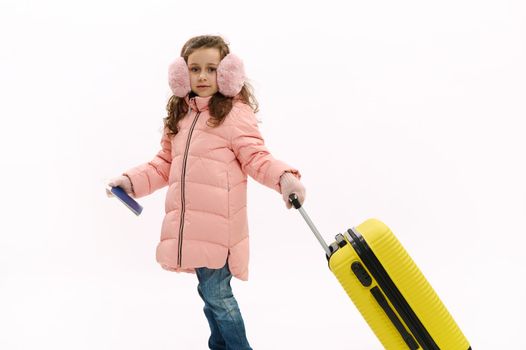 Fashionable little child, traveler girl in pink down coat, blue jeans and fluffy earmuffs, traveling abroad for winter holidays, walking with suitcase on white background. Journey Travel Tourism Trip