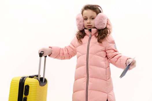 Caucasian adorable child, lovely little traveler girl with long curly hair, wearing pink fluffy earmuffs and down coat, posing with yellow suitcase and boarding pass over white background. Copy space