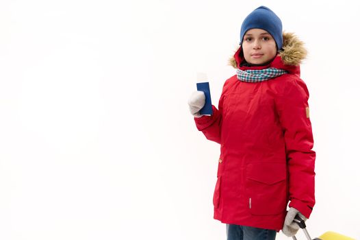 Adorable preteen boy wearing blue hat and red down parka, going for winter vacations, posing with boarding pass and suitcase, looking at camera, isolated over white background. Travel Journey Trip