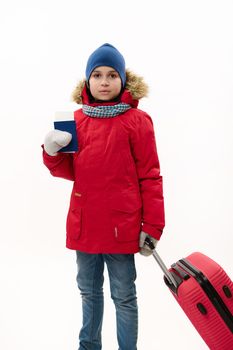 Full length portrait of teenage traveler boy wearing down jacket and blue jeans, going for winter vacations, walking with boarding pass and red suitcase over white background. Journey. Travel concept