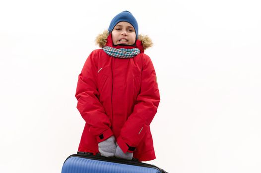 Middle-Eastern teenage boy, wearing red parka with fur hood, holding a blue suitcase, isolated over white background with free advertising space. Rest Getaway Journey Travel and Air flight concept