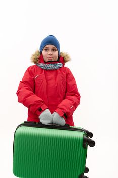 Happy multi-ethnic traveler child boy, wearing warm red down jacket, holding a green suitcase, going for winter vacations, isolated over white background with free advertising space. Kids travelling