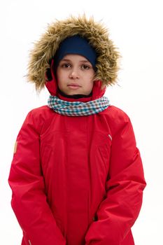 Confident vertical portrait of a handsome multi-ethnic child boy, wearing red down jacket with fur hood and blue scarf, looking at camera, isolated over white background. Cool weather. Winter concept