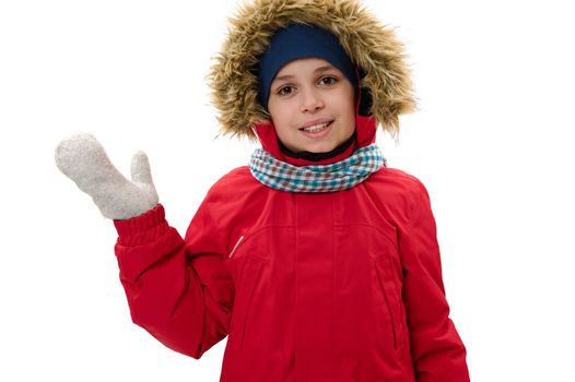 Adorable Caucasian teen boy in warm winter clothes, waves hello with hand, smiles a cheerful toothy smile, looking at camera, isolated on white background with copy space for your advertising text
