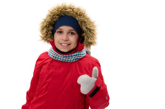 Caucasian cute teen boy in red down parka with fur hood and woolen mittens, thumbs up, smiles, looking at camera, isolated on white background with copy advertising space. Winter, cool weather concept
