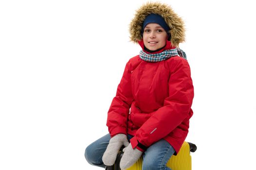 Isolated portrait on white background handsome child boy in warm red parka and blue denim jeans, sitting on a suitcase, smiling to camera. Free ad space. Winter travel. Journey. Rest getaway concept