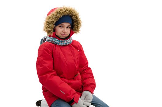 Portrait of multi-ethnic happy teenager traveler boy wearing red parka with fur hood, blue jeans and wool mittens, cutely smiling to camera, isolated over white background with free advertising space