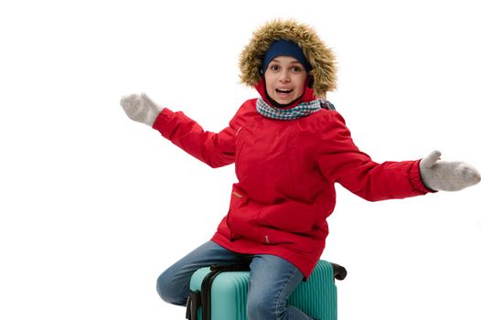 Cheerful boy in red parka with fur hood and woolen mittens, smiling with toothy smile, looking at camera, sitting on blue suitcase, traveling abroad on weekends or winter holidays, isolated on white