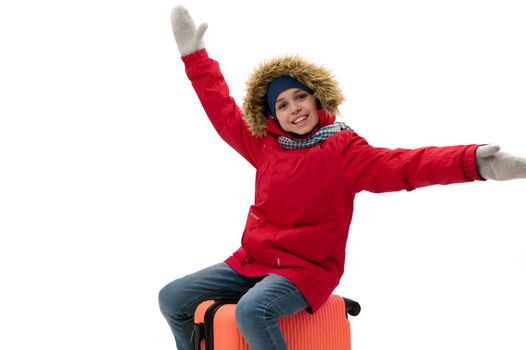 Caucasian smiling teenage traveler boy, wearing warm red down jacket, wool mittens and blue jeans, sitting on orange suitcase, travels abroad, isolated over white. Kids travelling on weekends getaway