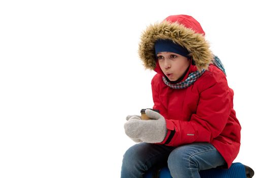 Charming preteen traveler boy in red down jacket and wool mittens, going for vacations, warms his hands with cup of hot drink, sits on suitcase, waits to board a flight. Travelling at winter holidays