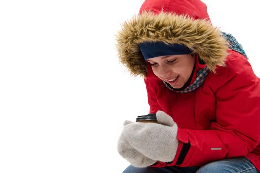 Preteen child boy, cute traveler adventurer in warm clothes, warming up, holding a disposable paper cup of hot drink, isolated over white background with free advertising space. Winter travel concept