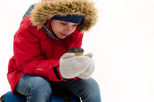 Adorable traveler teenage boy in red parka and jeans, sitting on blue suitcase, warming up with disposable cup of hot drink, isolated over white background. Advertising space. Winter travel concept