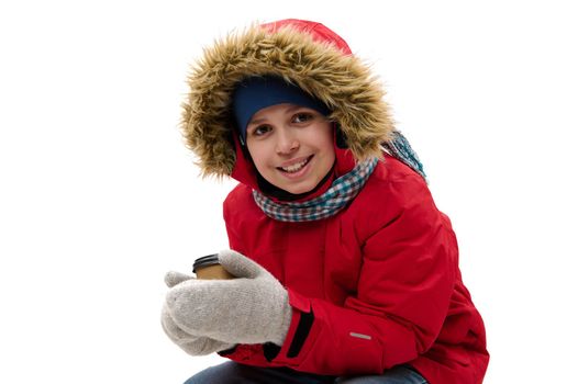Adorable multi-ethnic teenage boy in red parka with fur hood, blue scarf and wool mittens, holds a paper mug of hot drink, smiles to camera, isolated over white background with free advertising space