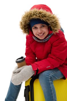 Happy traveler child boy in red down jacket, blue jeans, wool mittens smiling a toothy smile to camera, sitting on suitcase with a disposable cup of hot drink. Winter holidays. Travel. Journey concept