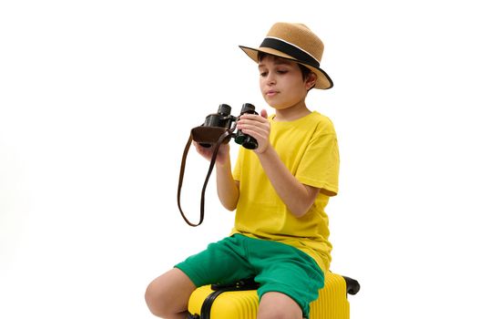 Delightful preteen child boy - young tourist traveler in yellow t-shirt, green summer shorts and straw hat, sitting on suitcase with binoculars in hands, isolated over white background. Free ad space