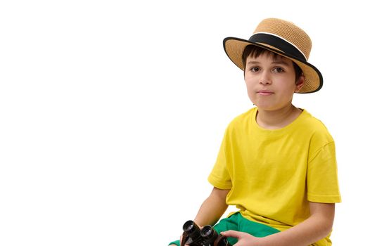 Close-up portrait of young tourist traveler, Caucasian teen boy in yellow t-shirt, green shorts and straw hat, sitting on suitcase, holding binoculars, looking at camera, isolated on white background