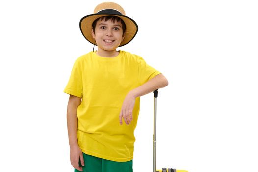 Caucasian joyful teen boy, tourist traveler in bright yellow t-shirt, green shorts and straw hat, smiling cheerful toothy smile looking at camera, posing with suitcase on white background. Copy space